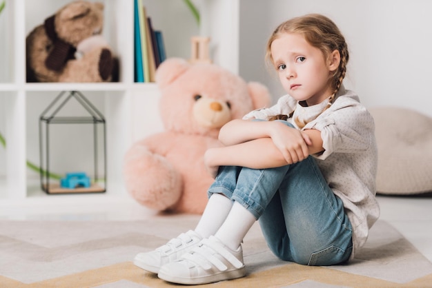 Lonely little child sitting on floor in front of shelves with toys and looking at camera