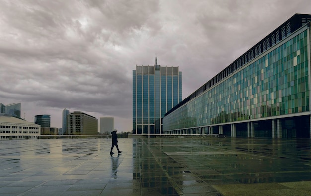 Lonely man in a city square on a rainy day. Cloudy urban landscape with reflections in the puddles o