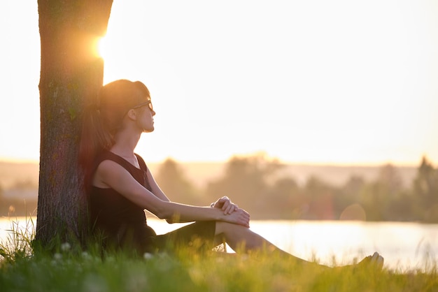 Lonely young woman sitting alone on grass lawn on lake shore enjoying warm evening Wellbeing and relaxing in nature concept