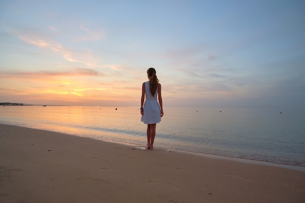 Lonely young woman standing on sandy beach by seaside enjoying warm tropical evening.