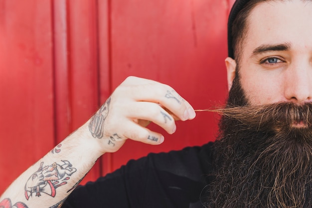 Long bearded young man pulling his mustache against wooden wall