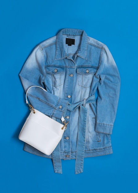 A long denim jacket and a white bag on a blue background Fashionable women's denim clothing