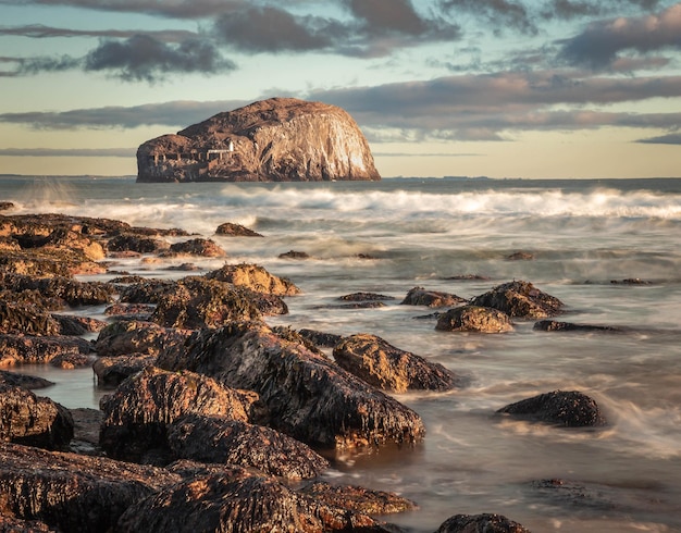 Long exposure image of Bass Rock Scotland from a rocky shore with sun shining on breaking waves