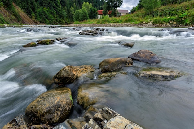 Long exposure of a mountain river with rocks from the bottom of the shot with trees and a house in t...