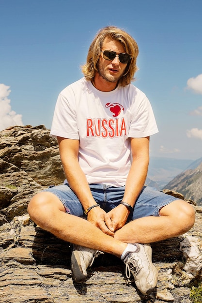 a long-haired guy in a white T-shirt with the inscription Russia on the background of mountains