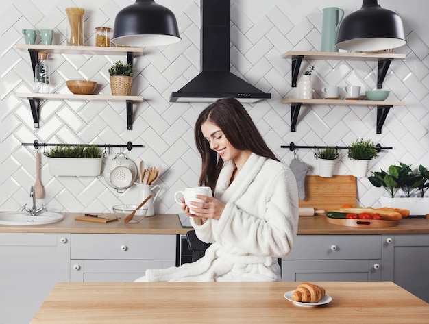 Long haired woman sitting with the cup of coffe at the light modern kitchen