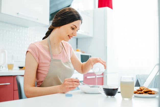 Long haired young woman wearing an apron sitting at the kitchen table and pouring milk into the bowl