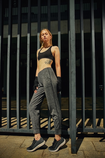 Long shot of beautiful sportive female in black hand warps who is posing by the metal fence