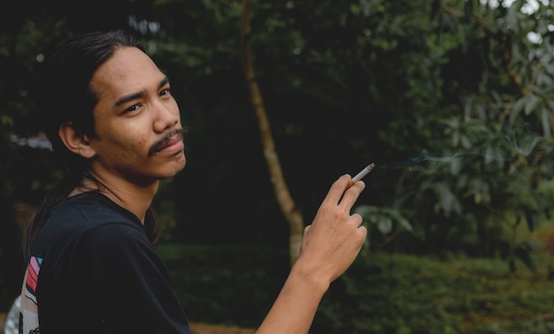 A longhaired Asian man with a beard holding a cigarette standing outside.