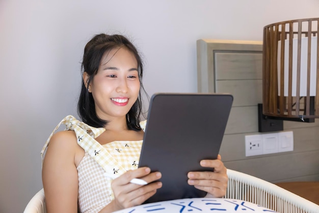 Longhaired Asian woman in a yellow dress is happily lounging on her tablet on the comfy sofa Online shopping social technology communication