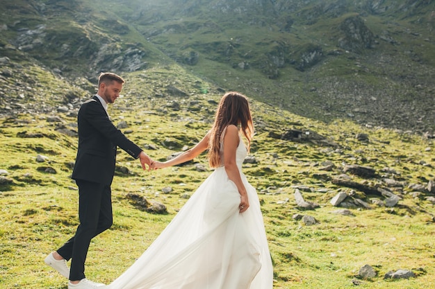 Longhaired bride in wedding dress and groom holding hands and walking in mountainside at sunset happ