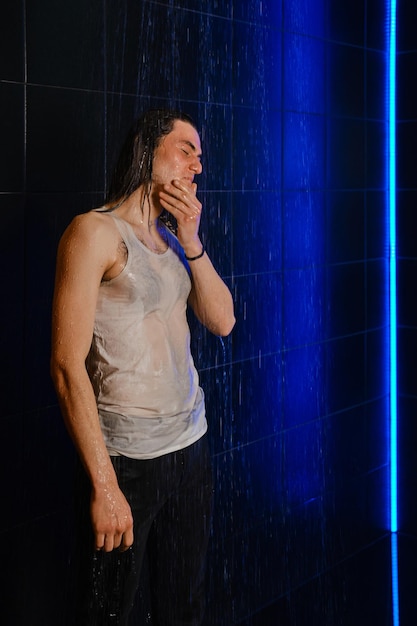 A longhaired man in wet tshirt stands in a shower illuminated by neon light