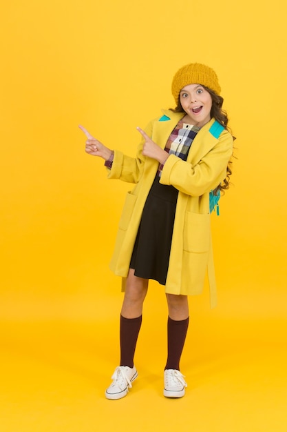 Look at closely Surprised child in autumn fashion pointing at something Small schoolgirl with open moth pointing yellow background Using pointing gesture to direct attention Pointing at product