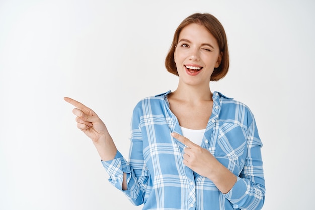 Look here. Smiling natural young woman winking, pointing fingers left at copy space, showing promo special deal, standing on white wall