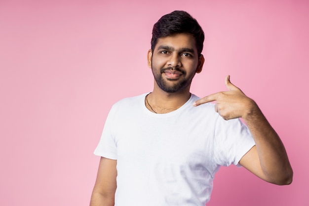 Look here. Waist up shot of indian man pointing at himself, showing blank space on white t shirt for your design, posing against pink background with copy space for your advertising.