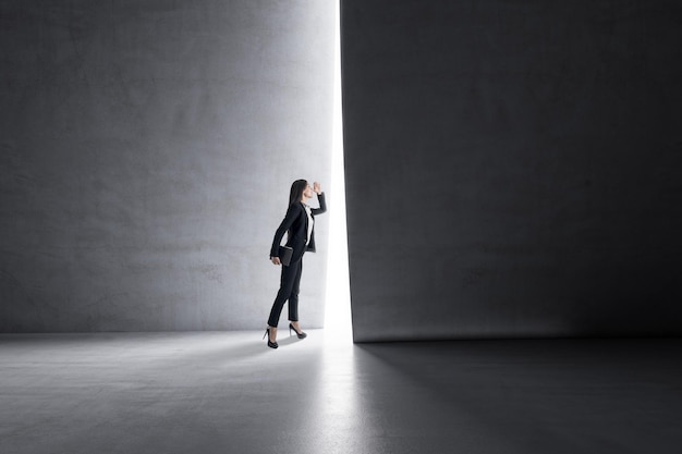 Look into the future and business success concept with businesswoman looking through a light hole in a dark wall in empty concrete space