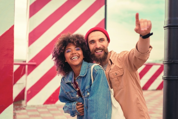 Look. Positive bearded young man smiling happily and pointing to the distance while hugging his cheerful girlfriend