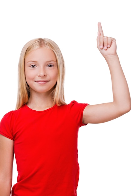 Look over there! Cheerful little girl pointing up and looking at camera while standing isolated on white