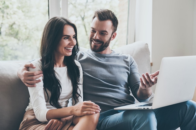 Look at this! Beautiful young loving couple looking at laptop and smiling while sitting together on the couch