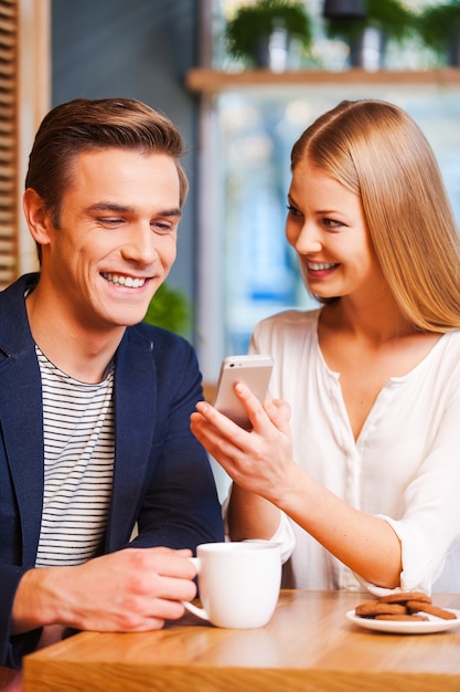 Look at this photo! Beautiful young woman showing something on mobile phone to her boyfriend while enjoying coffee in cafe together