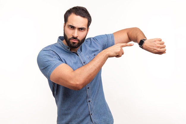 Look at time. Impatient bossy man with beard pointing finger at wrist watch and looking annoyed and displeased, showing clock to hurry up. Indoor studio shot isolated on white background