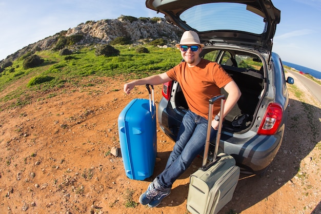 Looking at camera, a cheerful man going away for the weekend by the car with luggage.