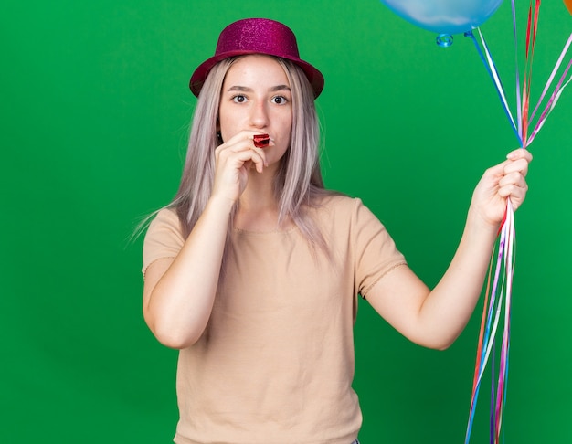 Looking at camera young beautiful girl wearing party hat holding party whistle in mouth 