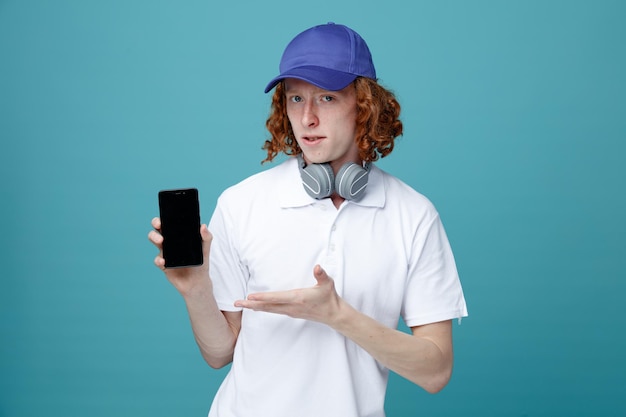 Looking at camera young handsome guy in cap wearing headphones holding and points at phone isolated on blue background