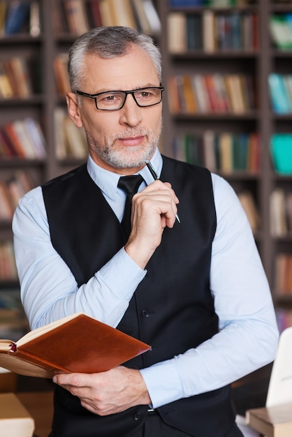 Looking for inspiration. Thoughtful grey hair senior man in formalwear holding note pad and looking away while leaning at the table and with bookshelf in the background