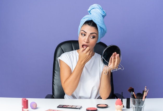 Looking at mirror beautiful woman sits at table with makeup tools wrapped hair in towel applying lipstick 