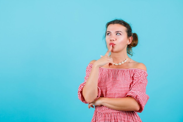 Looking up girl is thinking by holding forefinger on lips on blue background