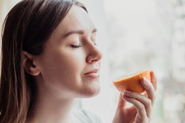 Loss of smell concept. Close up portrait of Caucasian young woman stands near the window and sniffs an orange