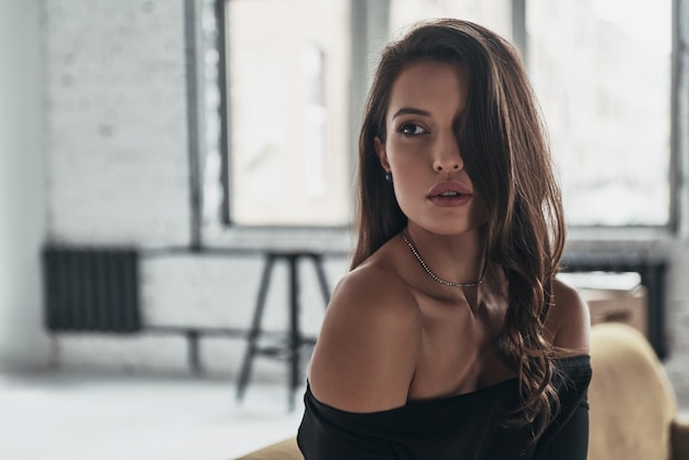 Lost in thoughts. Attractive young woman looking away while sitting on the sofa