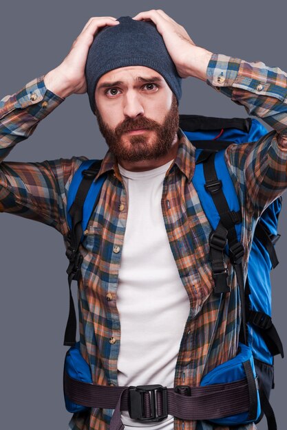 Lost tourist. Frustrated young bearded man carrying backpack and examining map while scratching head and standing against grey background