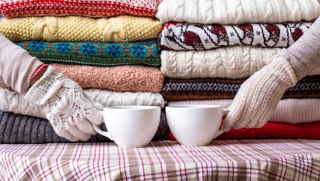 A Lot of Sweaters and Pullovers Different Colours folded in two Piles and hands with two cups of coffee.