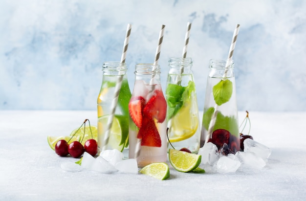 Lots of bottles with refreshing summer lemonade with lime, strawberry, cherry, cucumber and ice on a gray concrete background.