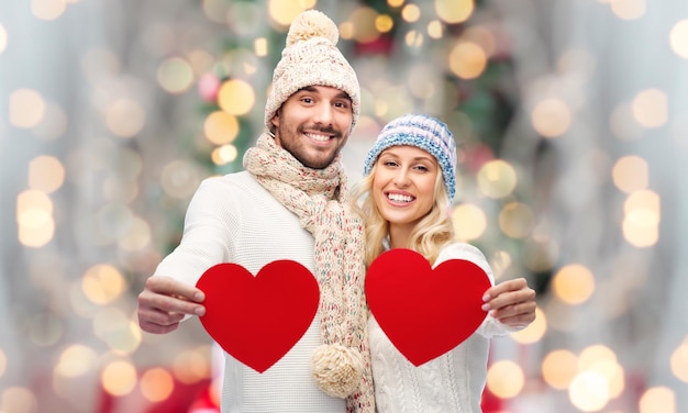 love, christmas, couple, holidays and people concept - smiling man and woman in winter hats and scarf holding red paper heart shapes over lights background