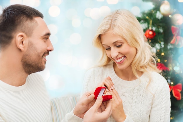 Photo love, christmas, couple, proposal and people concept - happy man giving diamond engagement ring in little red box to woman over blue holidays lights background