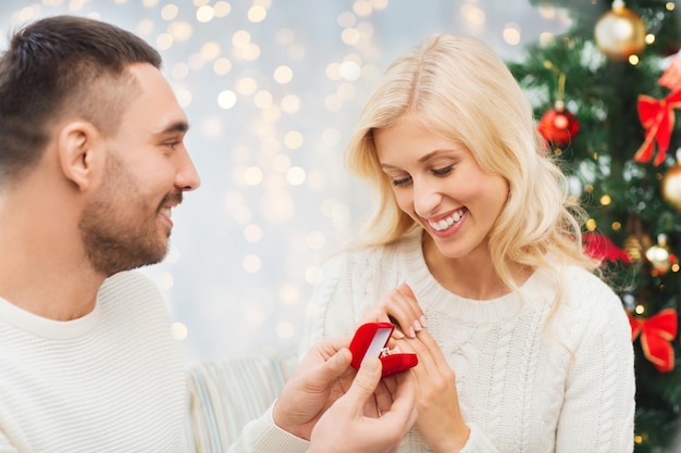 love, couple, proposal, holidays and people concept - happy man giving diamond engagement ring in little red box to woman over christmas tree and lights background