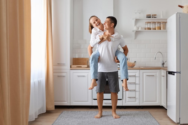 Love and family relationship Smiling young Caucasian couple in white shirts having fun husband holds wife on his back in cozy kitchen interior Male and female enjoy free time together at home