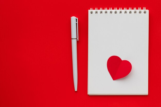 Love heart on notepad with pen on bright red background. Valentine's Day theme. Flat lay, top view, copy space.