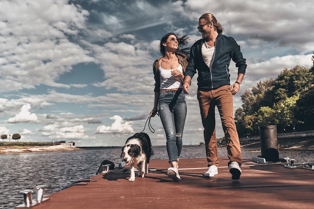 Love is in the air. Full length of beautiful young couple looking at each other and smiling while walking with their dog the river shore