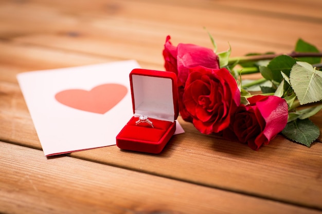 love, proposal, valentines day and holidays concept - close up of gift box with diamond engagement ring, red roses and greeting card on wood