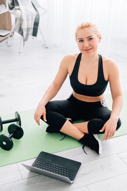 Love sport Happy athletic woman Online training Healthy body Inspired lady in black sportswear sitting lotus pose with dumbbells and laptop on floor in light home interior