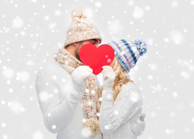 love, valentines day, couple, christmas and people concept - smiling man and woman in winter hats and scarf hiding behind red paper heart shape