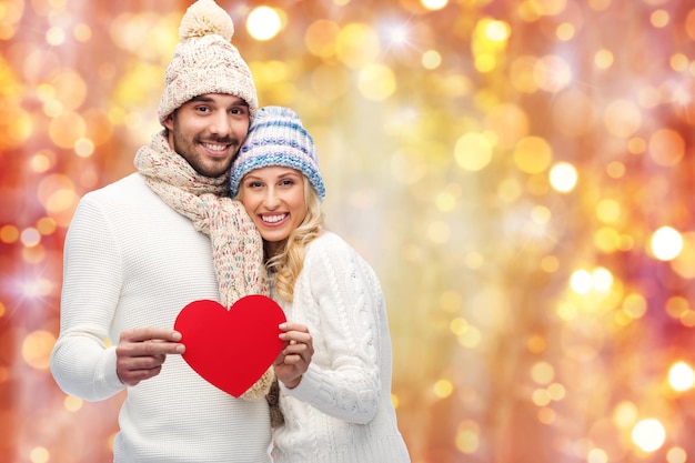 love, valentines day, couple, christmas and people concept - smiling man and woman in winter hats and scarf holding red paper heart shape over lights background