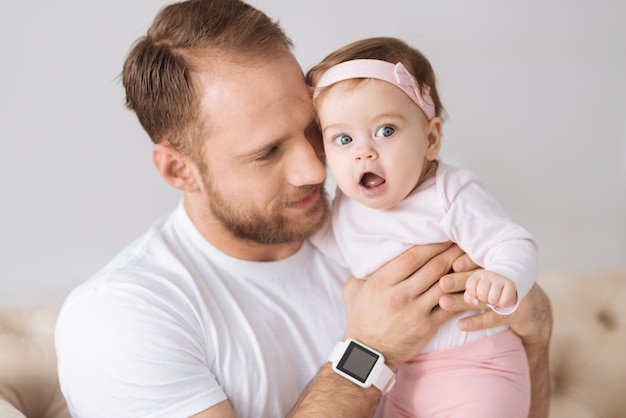 Love you. Charming positive young father sitting on the settee at home and hugging his toddler daughter while expressing positivity and love