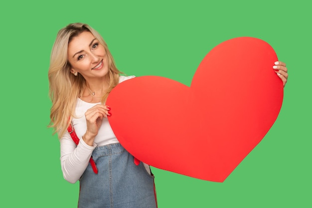 Love you, happy Valentine's day. Portrait of attractive adult blond woman holding red heart and smiling playfully, flirting, romantic congratulation. indoor studio shot isolated on green background