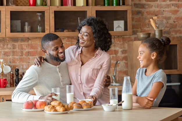 Lovely black family of three having lunch at kitchen, drinking milk and eating pastry, copy space