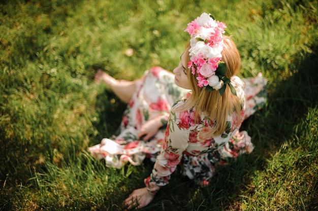 Lovely blonde woman dressed in flower dress and wreath on her head sitting on the green lawn